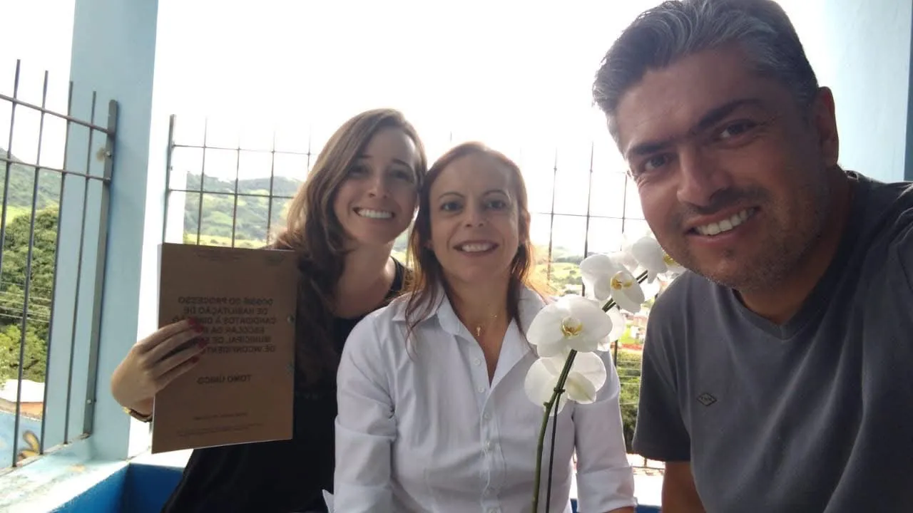 Duas mulheres e um homem sorrindo numa varanda; uma delas segura o dossiê encadernado.