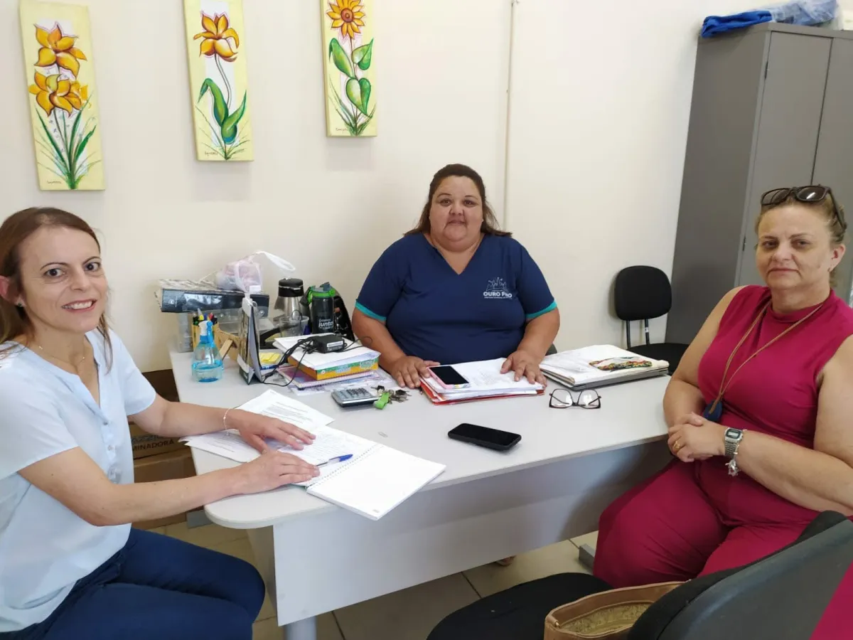 Três mulheres reunidas em volta de uma mesa de escritório com papéis e cadernos.