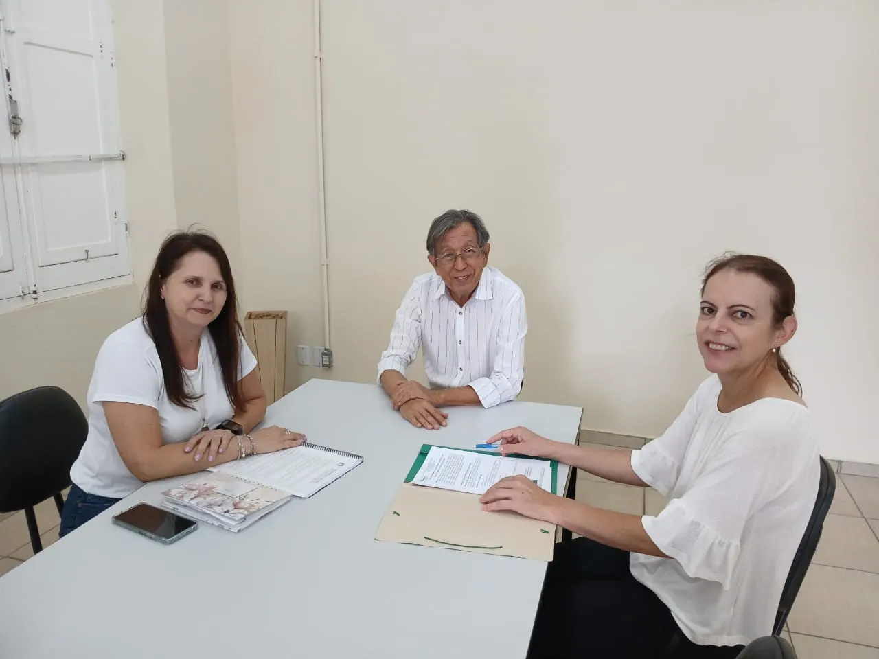 Duas mulheres e um homem sentados em volta de uma mesa, com pastas e documentos.