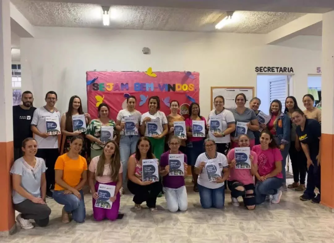 Grupo de professoras e professores de uma escola, diante do mural "Sejam bem-vindos", cada um segurando a apostila de Educação Infantil.